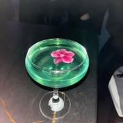 A coupe glass of pale green drink with a pink flower floating on top, on a dark marble table.