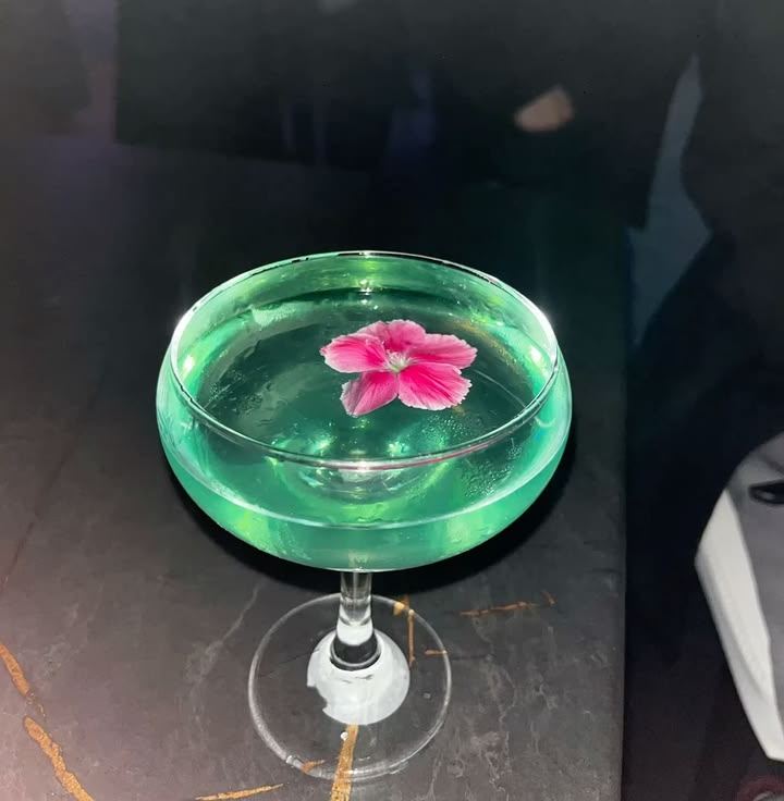 A coupe glass of pale green drink with a pink flower floating on top, on a dark marble table.