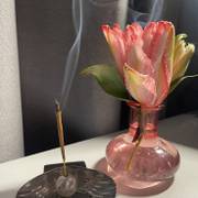 A pink parrot tulip in a pink glass vase beside an incense stick with rising smoke.
