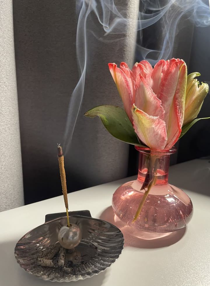 A pink parrot tulip in a pink glass vase beside an incense stick with rising smoke.