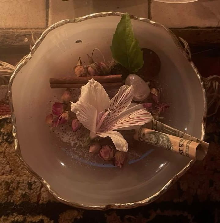 A pale shell-edged bowl holding dried rosebuds, a white lily, a cinnamon stick and a clear crystal.