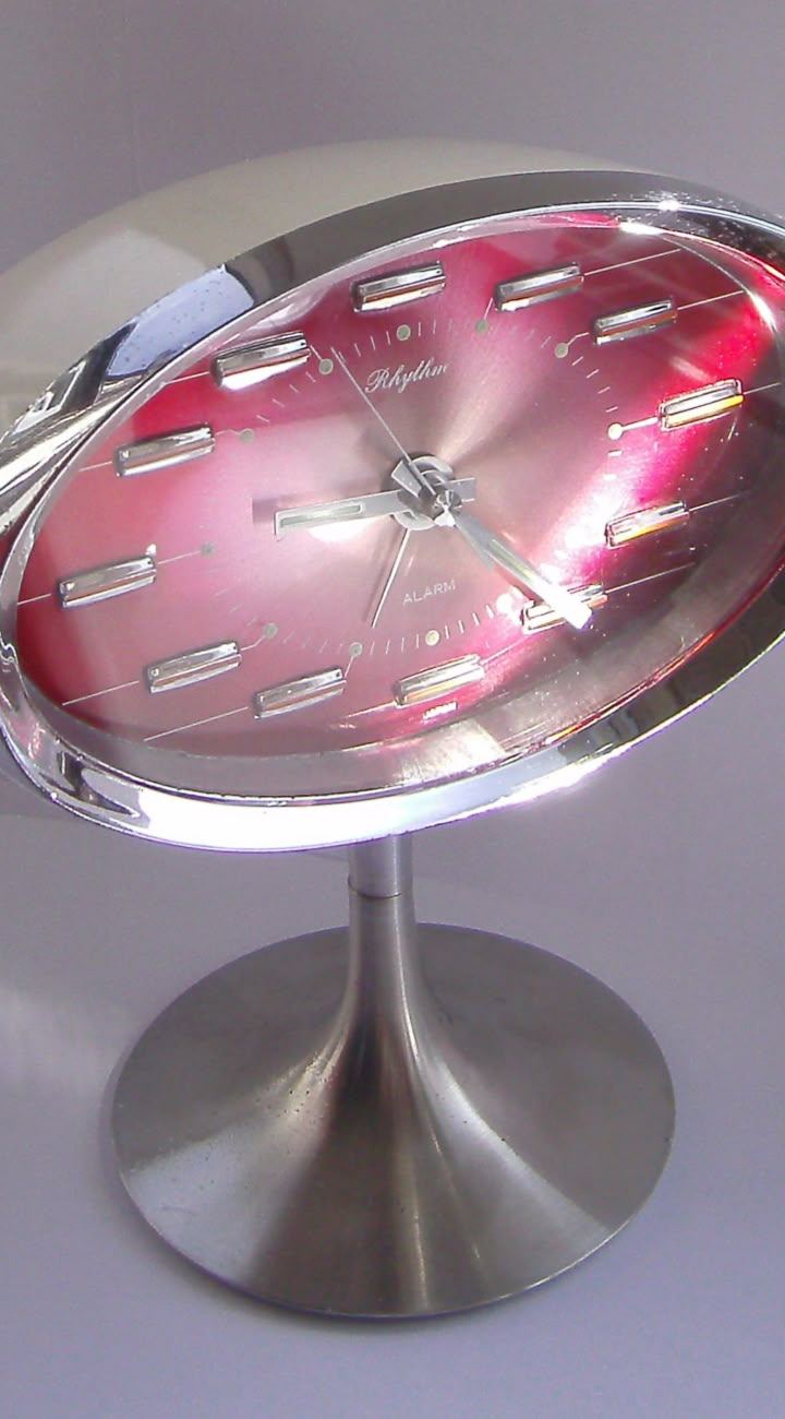 An oval clock with a pink face and reflective silver frame on a pedestal stand.